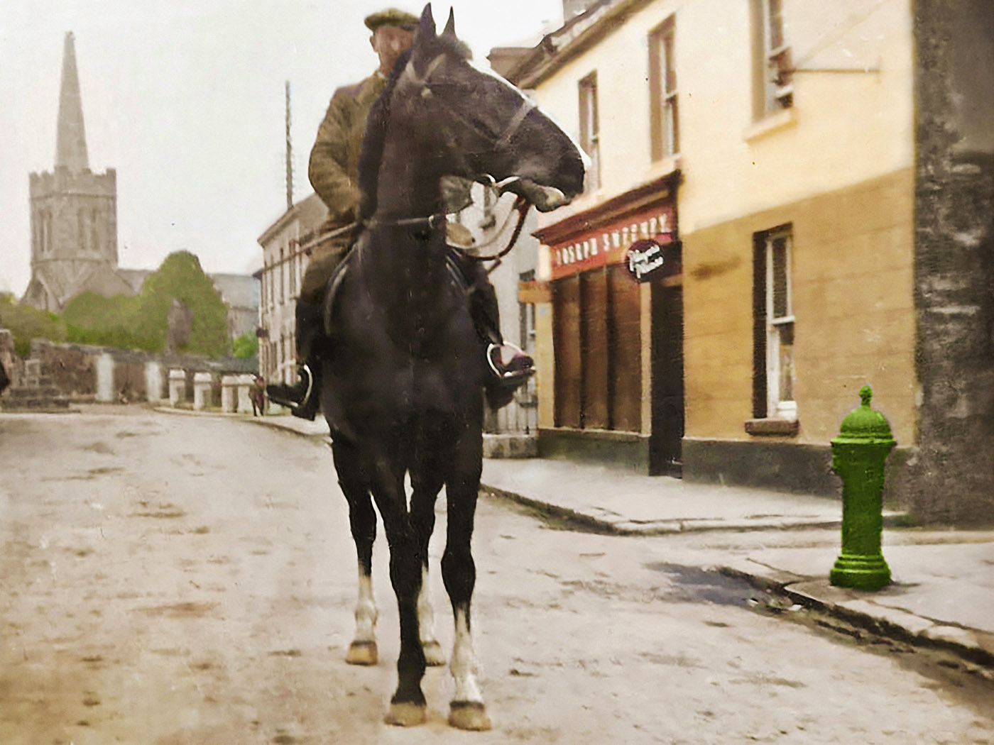 Photograph of Margie Murphy behind the mineral bar which she ran for decades in The Town Hall, Athenry.  She’s accompanied by Tom Fahy who also ran shops in the town including Fahy’s VG Foodmarket on Northgate Street.  Behind them are ads for Fox’s Glacier mints, Afton Cigarettes, Aspro, Cadbury’s Choffees, some Tayto Quips cards, posters for several showbands including the Athenry band Premier Aces. 