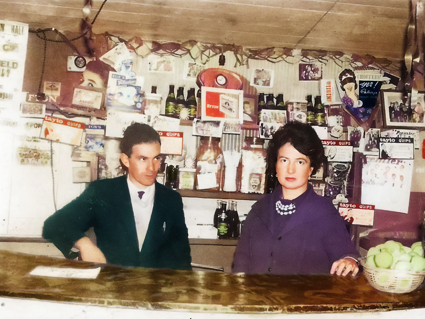 Photograph of Margie Murphy behind the mineral bar which she ran for decades in The Town Hall, Athenry.  She’s accompanied by Tom Fahy who also ran shops in the town including Fahy’s VG Foodmarket on Northgate Street.  Behind them are ads for Fox’s Glacier mints, Afton Cigarettes, Aspro, Cadbury’s Choffees, some Tayto Quips cards, posters for several showbands including the Athenry band Premier Aces. 