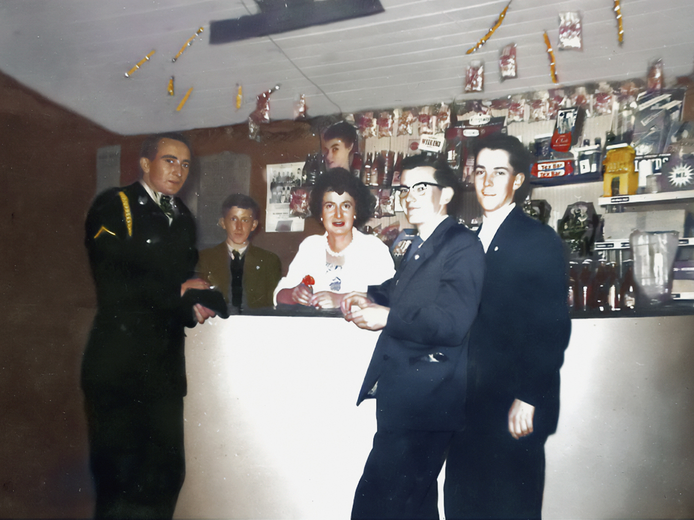 Photograph of Margie Murphy behind the mineral bar which she ran for decades in The Town Hall, Athenry.  She’s accompanied by Tom Fahy who also ran shops in the town including Fahy’s VG Foodmarket on Northgate Street.  Behind them are ads for Fox’s Glacier mints, Afton Cigarettes, Aspro, Cadbury’s Choffees, some Tayto Quips cards, posters for several showbands including the Athenry band Premier Aces. 